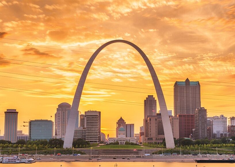 Gateway Arch with downtown St. Louis skyline at sunset and an orange sky over the river.